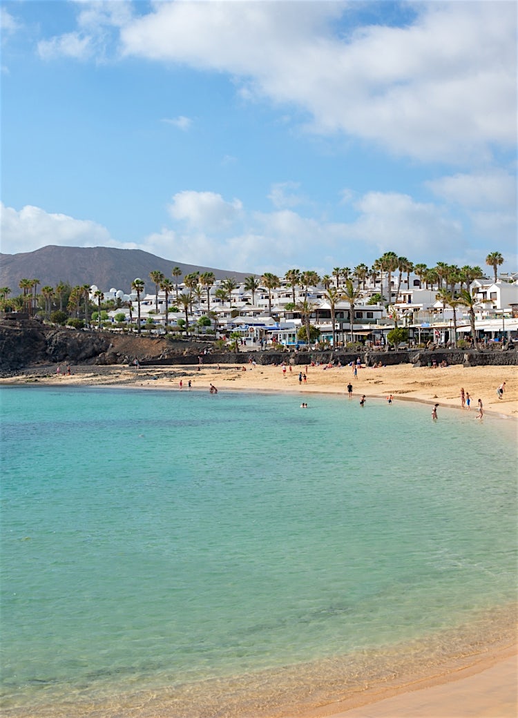 Turquoise-water beach in Lanzarote with palm-lined waterfront, whitewashed buildings and volcanic mountains in the background.