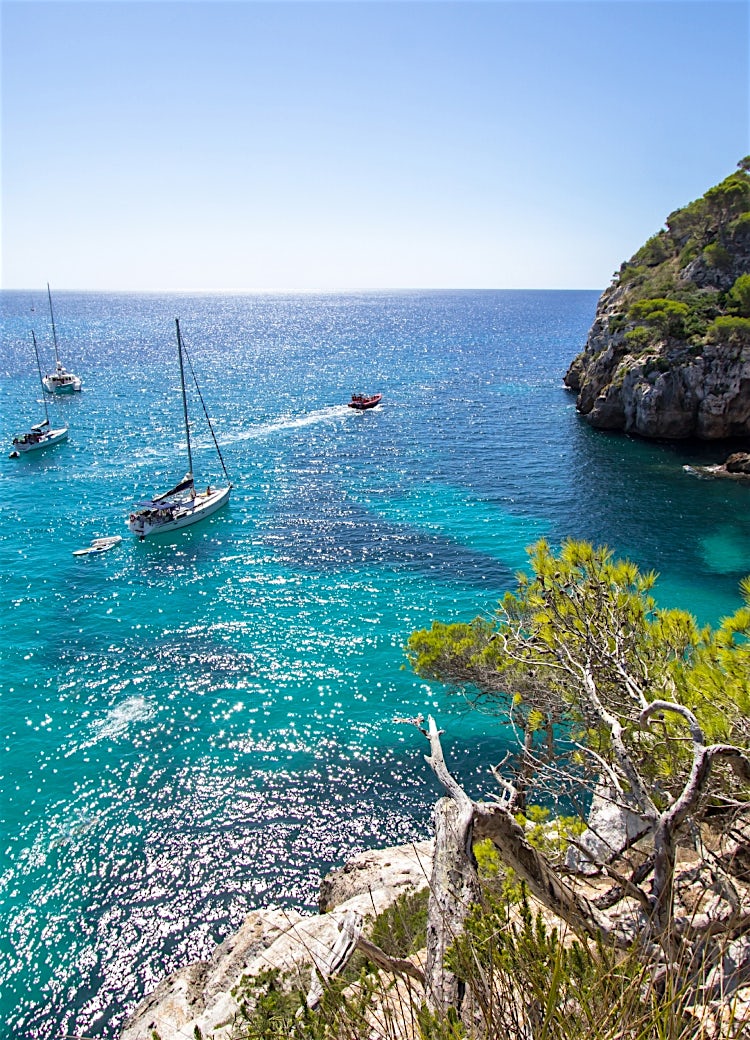 Menorca turquoise cove with sailboats, rocky cliffs and crystal-clear Mediterranean waters, Balearic Islands, Spain.