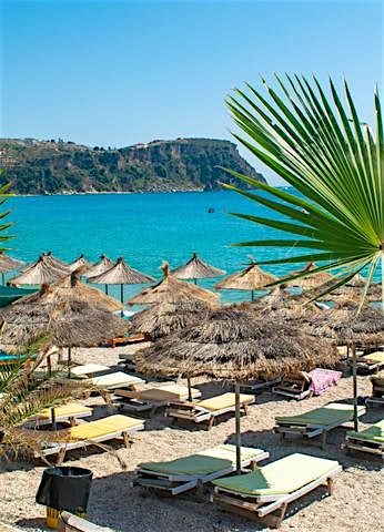 Albanian Riviera beach with turquoise Ionian Sea, palm trees, straw parasols, sun loungers, and scenic coastal resort views.