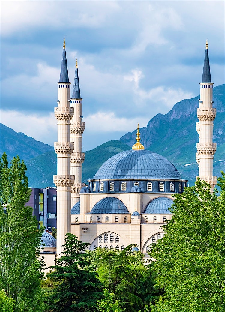 Grand mosque in Albania with towering minarets, domed architecture, lush greenery, and dramatic mountain scenery in the background.