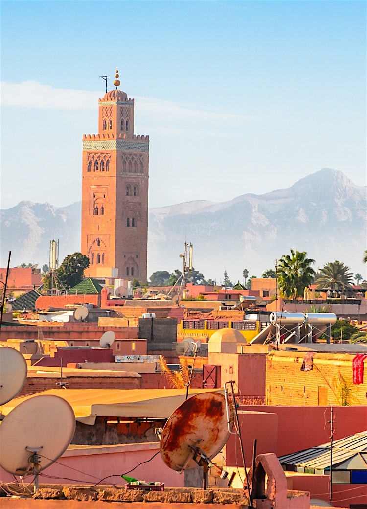 Marrakech skyline with the Koutoubia Mosque, traditional Moroccan architecture, palm trees, and Atlas Mountains in the background.