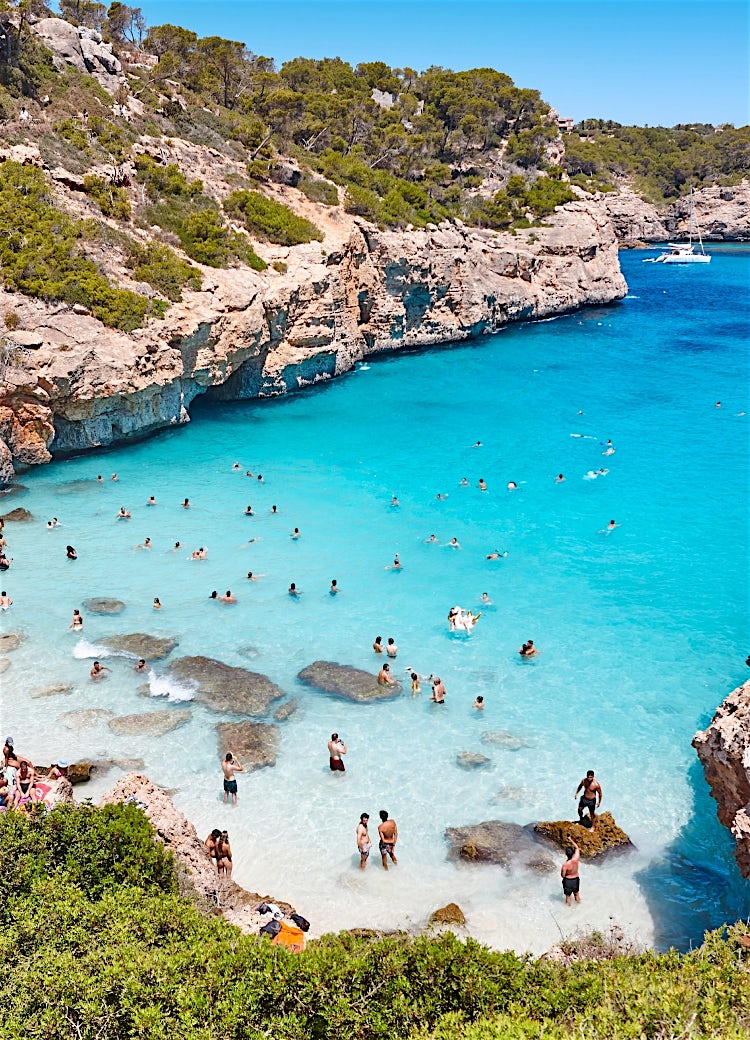 Balearic Islands turquoise cove with crystal-clear waters, rocky cliffs, swimmers, anchored yachts and stunning Mediterranean coastal scenery.