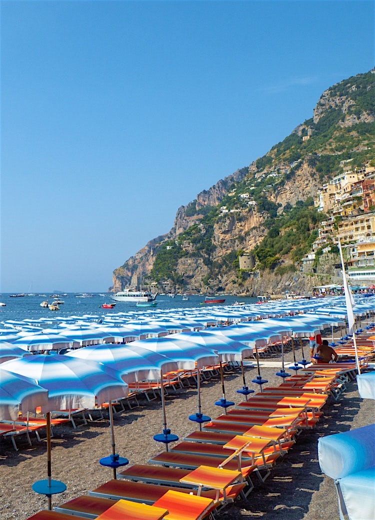 Amalfi Coast beach with blue-striped umbrellas, Mediterranean waters, luxury boats and Positano’s cliffside village backdrop.