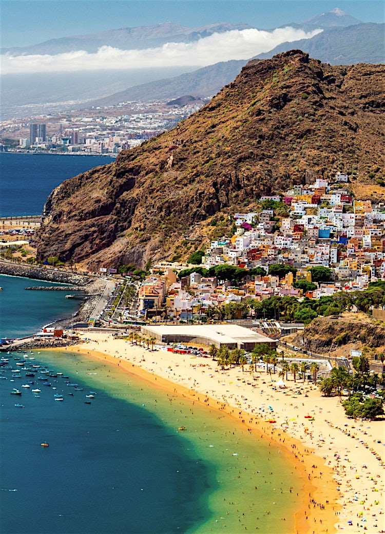 Canary Islands coastline with colourful hillside village, sandy beach, turquoise Atlantic Ocean, marina, and dramatic volcanic mountain scenery.
