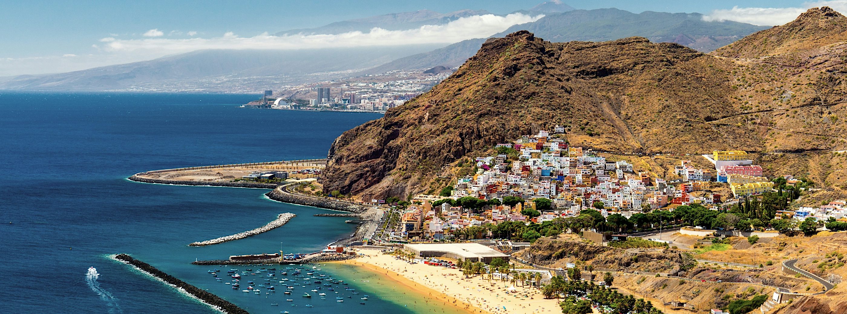 Canary Islands coastline with colourful hillside village, sandy beach, turquoise Atlantic Ocean, marina, and dramatic volcanic mountain scenery.