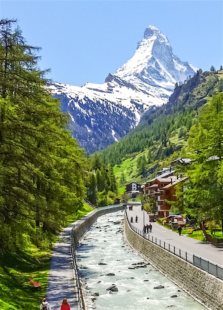 Zermatt, Switzerland, with alpine chalets, mountain stream, lush valley landscape, and the iconic Matterhorn peak in the background.