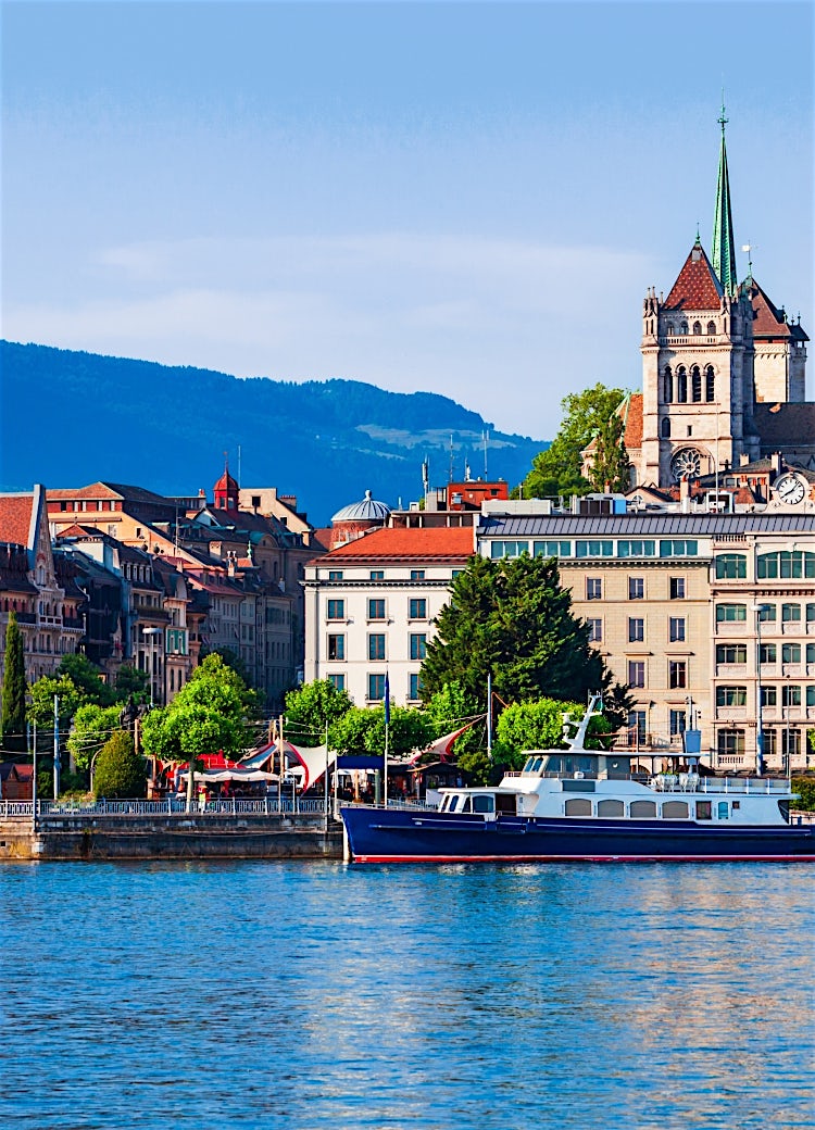 Geneva, Switzerland skyline with lakeside ferris wheel, historic city architecture, and St. Pierre Cathedral against a mountain backdrop.