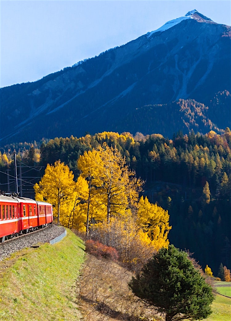 Scenic Swiss mountain railway with a red train travelling through alpine countryside, surrounded by forests, valleys, and snow-capped peaks.