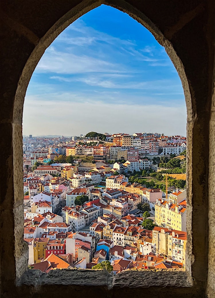 Panoramic view of Lisbon, Portugal, featuring colourful hillside buildings, red-tiled rooftops and the historic cityscape framed by stone architecture.
