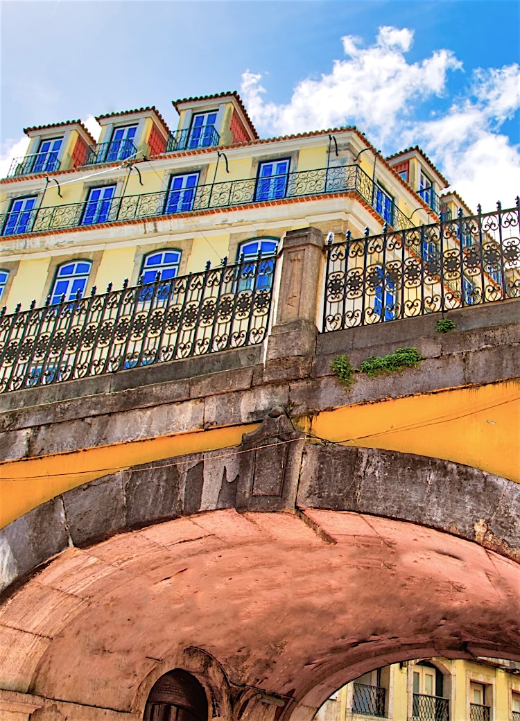 Historic Lisbon architecture featuring a traditional archway, ornate iron railings and colourful Portuguese buildings under a blue sky.