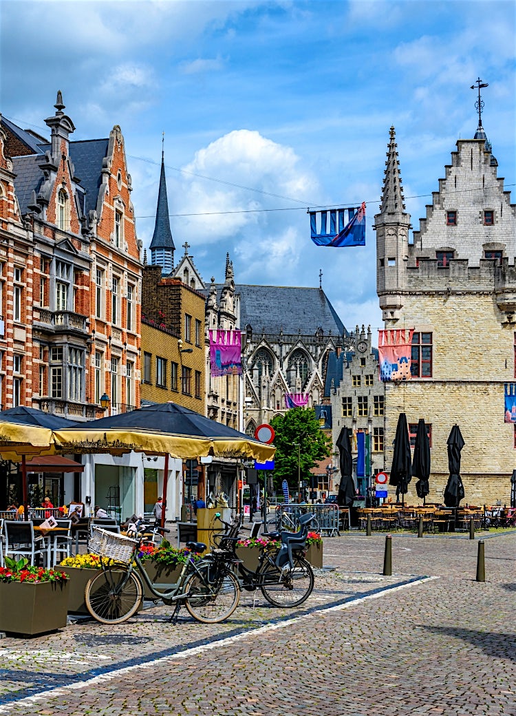 Historic Belgian city square with medieval stone guildhall, Flemish architecture, outdoor cafés, colorful banners and Gothic church in Belgium.