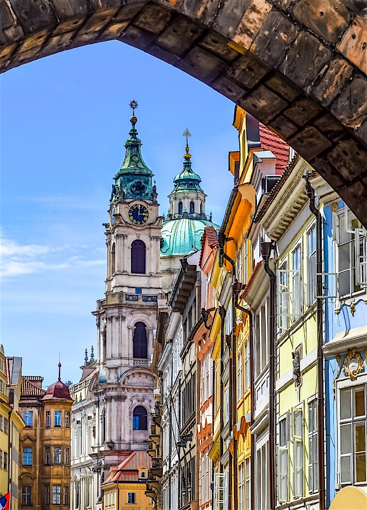 Historic Prague Old Town street framed by a stone arch, featuring colorful Baroque buildings and St. Nicholas Church clock tower under a bright blue sky.