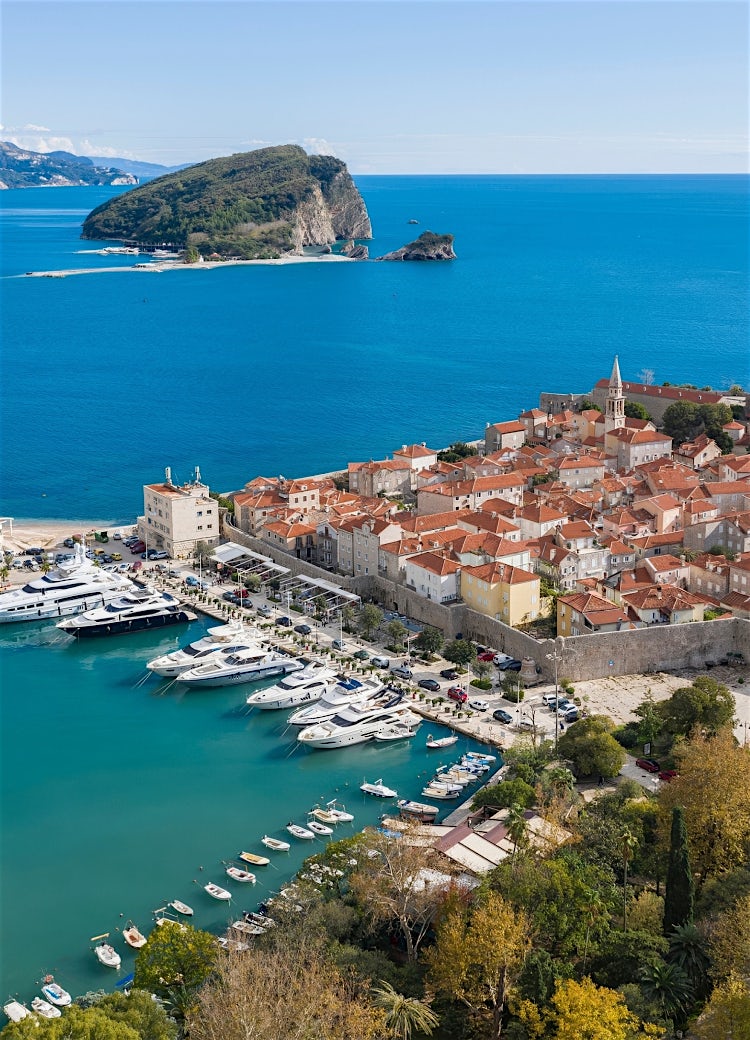 Panoramic view of Budva Old Town, Montenegro, with red-tiled rooftops, Adriatic Sea, marina, and Sveti Nikola Island under a clear blue sky.