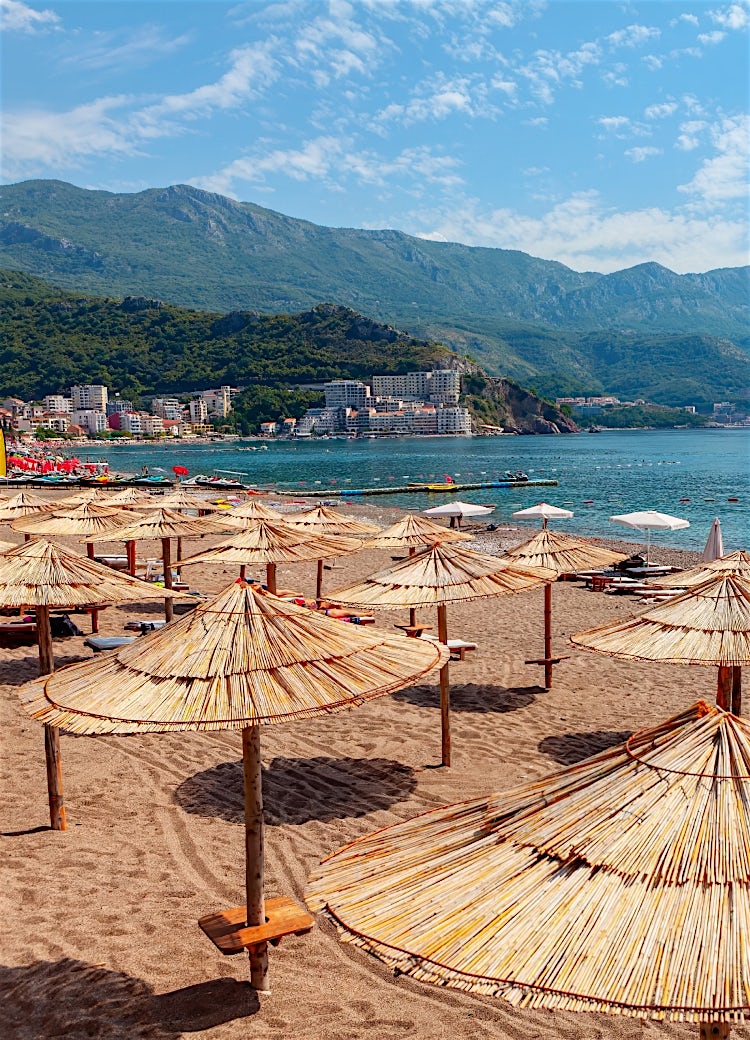 Montenegro beach with straw parasols, Adriatic coastline, seaside resort hotels, and mountain backdrop under a sunny sky.