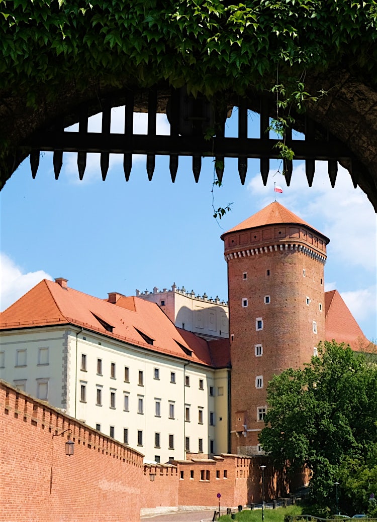 Wawel Royal Castle in Kraków, Poland, viewed through a historic stone archway with lush greenery and medieval red-brick towers.