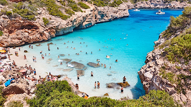 Balearic Islands turquoise cove beach with swimmers, crystal-clear water, rocky cliffs, secluded bay and Mediterranean coastal scenery.