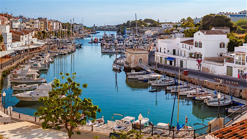 Menorca marina with yachts, waterfront promenade, historic harbor buildings and scenic Mediterranean coastal town views.