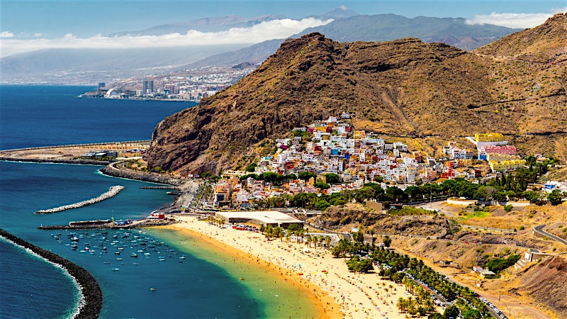 Canary Islands coastal resort with golden beach, turquoise bay, colorful hillside village and dramatic volcanic mountain scenery.