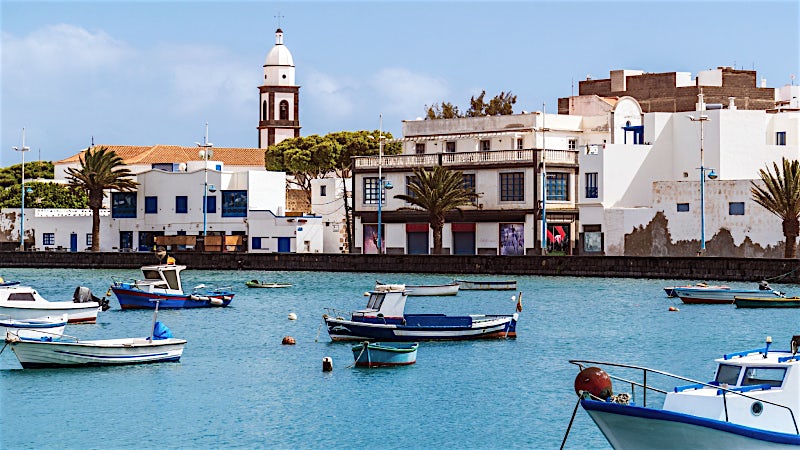 Lanzarote waterfront with fishing boats, traditional whitewashed buildings, church tower and scenic harbor views.