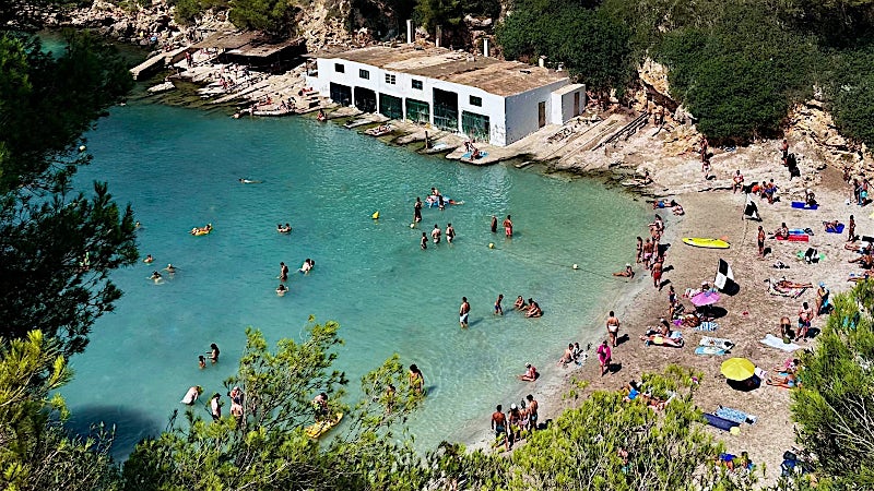Turquoise cove beach in Majorca, Spain, with swimmers, sunbathers, crystal-clear water, rocky coastline and waterfront beach club.