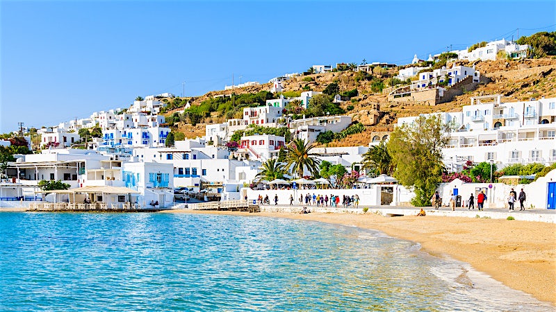 Mykonos Town waterfront with whitewashed Cycladic houses, sandy beach and Aegean Sea, Greece.