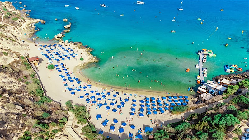 Aerial view of Konnos Bay with crystal-clear water, sandy cove and blue beach umbrellas, Cyprus.