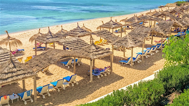 Sandy Tunisia beach with sun loungers, thatched parasols and clear Mediterranean waters.