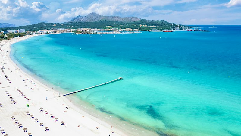 Aerial view of Mallorca beach with turquoise Mediterranean sea, white sand coastline and mountain backdrop.