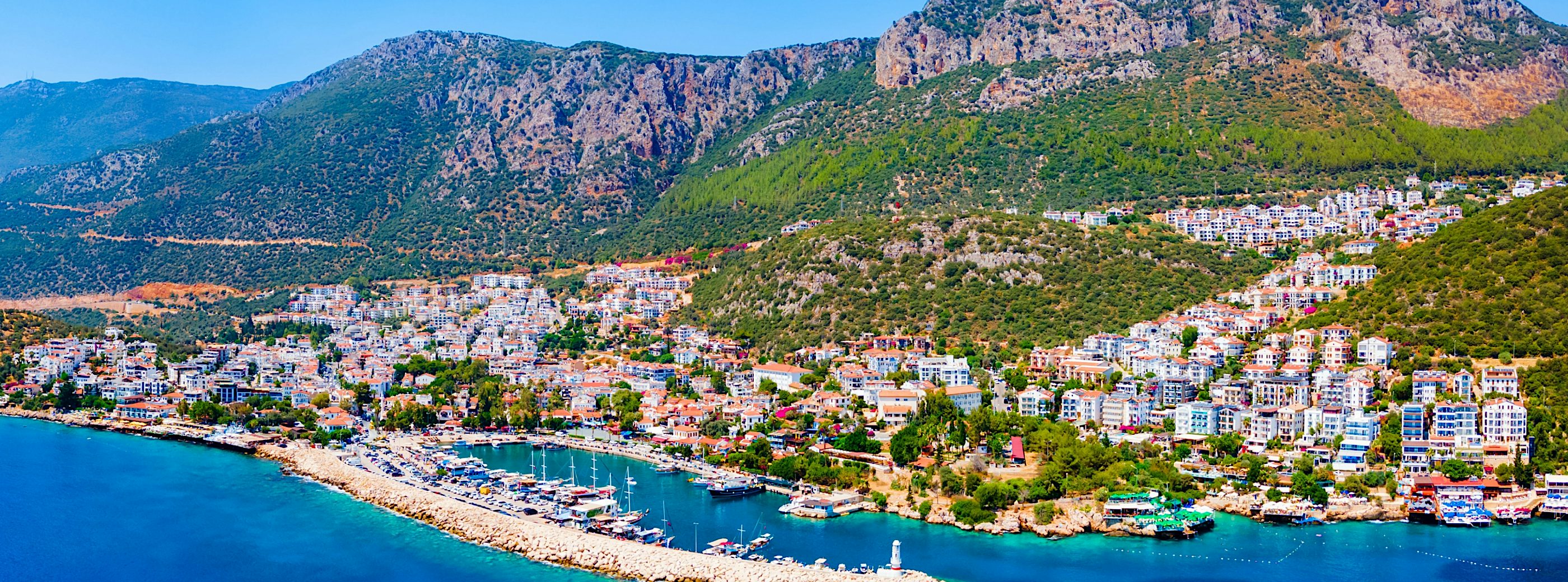 Aerial view of a Turkish Riviera coastal town with marina, turquoise Mediterranean waters, hillside homes, and rugged mountain backdrop under clear blue skies.