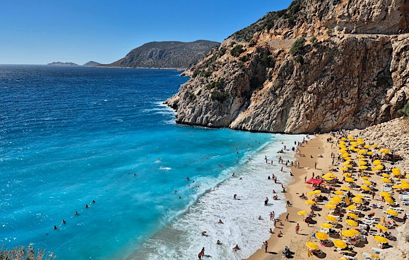 Turquoise sea laps a sandy beach beneath rugged cliffs, with swimmers and yellow parasols, ideal for value holidays in the sun.